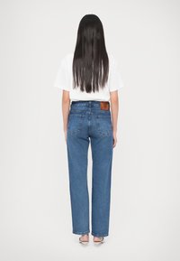 Femme aux longs cheveux foncés portant un t-shirt blanc, un jean bleu taille haute à jambes droites et des talons beiges, debout de dos.