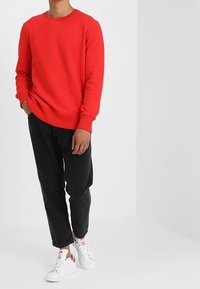 Persona vistiendo un sudadera roja brillante, jeans negros y zapatillas blancas, de pie con una mano en el bolsillo contra un fondo claro y sencillo.