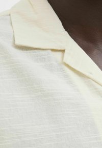 Camisa de color beige claro con un tejido texturizado. Presenta un cuello con solapa y sutiles rayas horizontales. Material suave y transpirable.