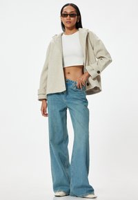 Beige kort jacka med huva, knapppdetaljer; vit kort topp; ljusblå vida jeans; modell bär svarta solglasögon.