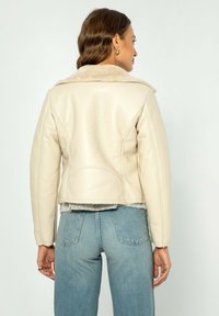Giacca di pelle color crema con un colletto peloso, design corto e texture liscia, abbinata a jeans azzurri e dettagli di cucitura visibili.