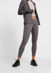 Šedá sportovní bunda s texturovanými rukávy, spárovaná s odpovídajícími přiléhavými legínami s síťovými detaily. Černé sportovní boty dotvářejí celý outfit.