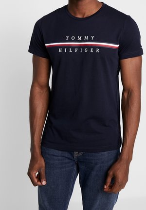 T-shirt en coton bleu marine avec col rond, manches courtes et motif à rayures horizontales rouges et blanches. Le nom de la marque "TOMMY HILFIGER" est centré.