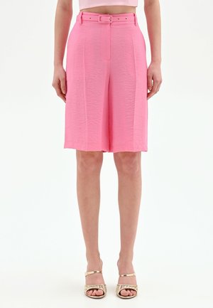 Donna che indossa pantaloncini rosa a vita alta con cintura fino al ginocchio, abbinati a sandali con tacco rigati e punta aperta, in piedi su sfondo bianco.