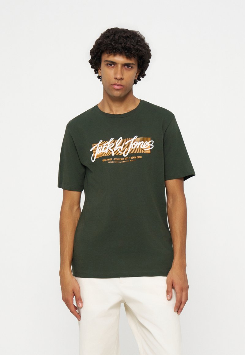 Joven con cabello rizado que lleva una camiseta verde oscuro con texto blanco y naranja y pantalones de color claro, de pie frente a un fondo blanco liso.