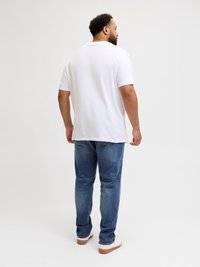 Λευκό κοντομάνικο μπλουζάκι, σε χαλαρή γραμμή, συνδυασμένο με μπλε τζιν παντελόνι και λευκά αθλητικά παπούτσια με καφέ σόλα. Το φόντο είναι απλό.