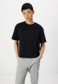 Camiseta negra de algodón con cuello redondo y mangas cortas, combinada con pantalones gris claro, de pie contra un fondo blanco.