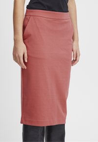 Jupe mi-longue ajustée en tissu doux de couleur rose pâle. Elle présente une large taille et des poches latérales, avec une texture lisse et des lignes épurées.