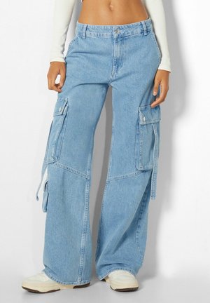 Vrouw draagt lichtblauwe wijde denim cargobroek met grote klepzakken en witte sneakers, staand tegen een effen achtergrond.