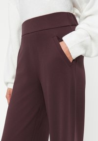 Pantalones de pierna ancha burdeos con una cintura alta y bolsillo lateral. Tejido suave con una textura lisa. Combinados con un suéter blanco.