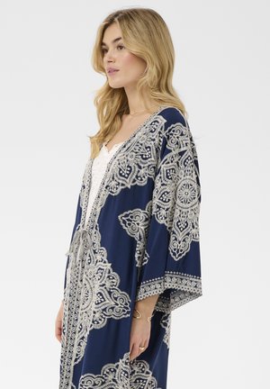 Femme blonde portant un kimono bleu marine orné de motifs blancs complexes semblables à de la dentelle, sur un haut blanc, debout devant un fond uni.