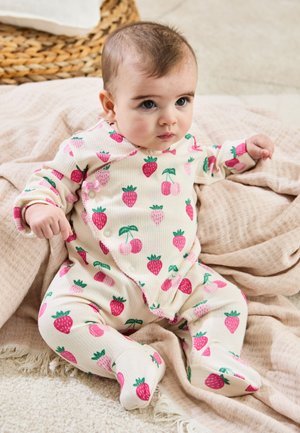 Baby zit op een beige deken, draagt een wit rompertje met roze aardbei- en kersenpatronen en kijkt aandachtig opzij.