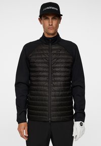 En man står självsäkert iklädd en svart isolerad jacka med dragkedja fram och en keps, hållande en golfhandske i sin vänstra hand.