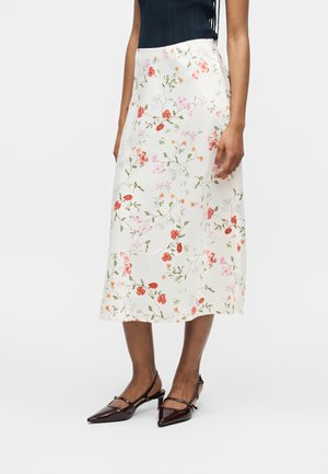 Frau trägt einen weißen Midi-Rock mit rosa und roten Blumenmustern, kombiniert mit dunkelspitzen Slingback-Pumps und einem schwarzen ärmellosen Top.