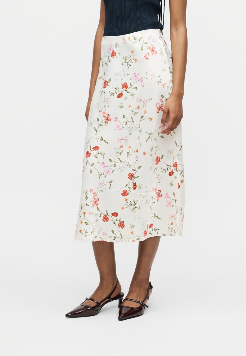 Γυναίκα φοράει μια λευκή midi φούστα με ροζ και κόκκινα λουλουδάτα σχέδια, συνδυασμένη με σκουρογάλαζα μυτερά πέδιλα με λουράκι και μαύρο αμάνικο τοπ.