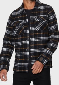 Chemise en flanelle noire à carreaux avec des lignes blanches et orange, fermeture à boutons, deux poches poitrine et un col classique.