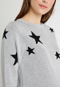 Sweater de malha cinza com padrões de estrelas pretas. Gola redonda e mangas longas, com uma textura suave e design casual.