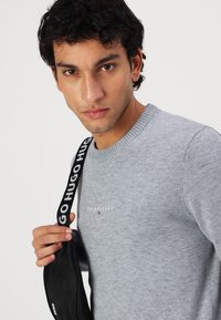 Szary sweter z okrągłym dekoltem i prążkowanym wykończeniem, z logo "TOMMY JEANS". Model trzyma czarną torbę z paskiem oznaczonym jako "HUGO."
