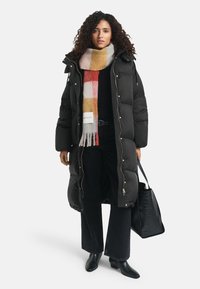 Vrouw met krullend haar, gekleed in een lange zwarte pufferjas, een meerkleurige geruite sjaal, zwarte broek, laarzen en een zwarte gestructureerde handtas.