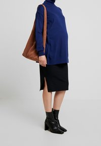 Marineblauer Rollkragenpullover, schwarzer Midirock mit seitlichem Schlitz und schwarze Stiefeletten. Eine braune Schultertasche ergänzt das Outfit.