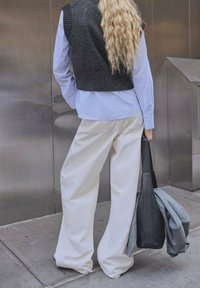 Schwarze Strickweste über einem hellblauen Hemd mit Knöpfen, kombiniert mit cremefarbenen, weit geschnittenen Hosen. Ein grauer Pullover und eine schwarze Tasche werden gehalten.