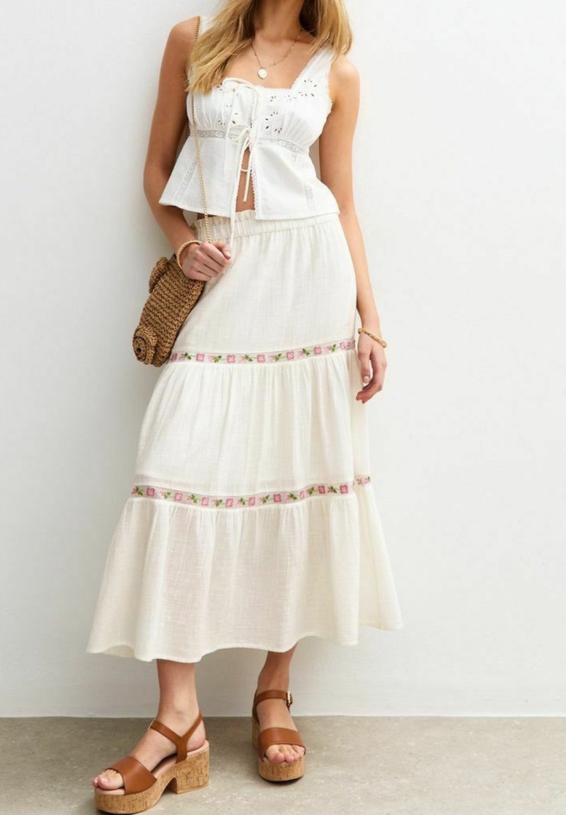 Blouse en coton blanc avec des détails en broderie, associée à une jupe maxi blanche à volants ornée de broderies florales roses. Des sandales à plateforme brunes complètent le look.