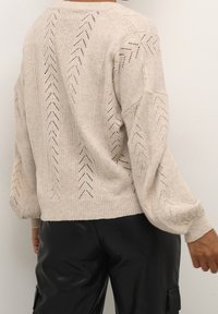 Béžový pletený svetr s volným střihem, s texturou ve tvaru šípů a vroubkovanými manžetami. Nositý přes černé kožené kalhoty.