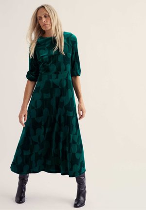 Femme aux longs cheveux blonds portant une robe midi verte à motifs et des bottes noires, debout devant un fond clair uni.