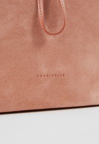 Sac à main en daim rose doux avec poignée en cuir et logo COCCINELLE embossé sur le devant, sur fond blanc.