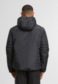 Chaqueta acolchada negra con capucha, que presenta un exterior suave y brillante, puños elásticos y un corte holgado. Sin logotipos visibles.
