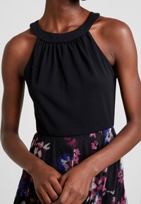 Černý halter top s nařasenými detaily kolem krku, spárovaný s květinovou, průhlednou sukní v odstínech růžové a modré.