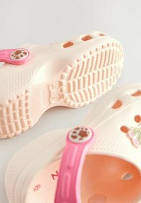 Rosa und cremefarbene Kunststoff-Clogs mit einer strukturierten Sohle, Belüftungslöchern und einem rosa Riemen mit einem kreisförmigen Tierdruckakzent.
