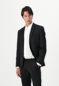 Veste de costume noire avec un revers en pointe, fermeture à un bouton et poches latérales, associée à une chemise blanche à boutons et un pantalon noir.
