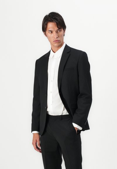 Veste de costume noire avec un revers en pointe, fermeture à un bouton et poches latérales, associée à une chemise blanche à boutons et un pantalon noir.