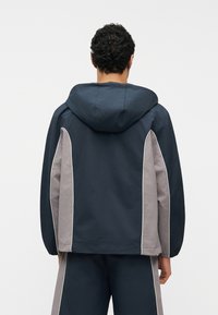 Άτομο που φοράει μπλε ναυτικό και γκρι μπουφάν με κουκούλα και ταιριαστό παντελόνι, εμφανιζόμενο από πίσω σε απλό λευκό φόντο.