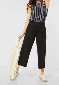 Kvinna iklädd svarta vida cropped-byxor, randig ärmlös topp, vita sneakers, håller en ljus denimjacka i ena handen.