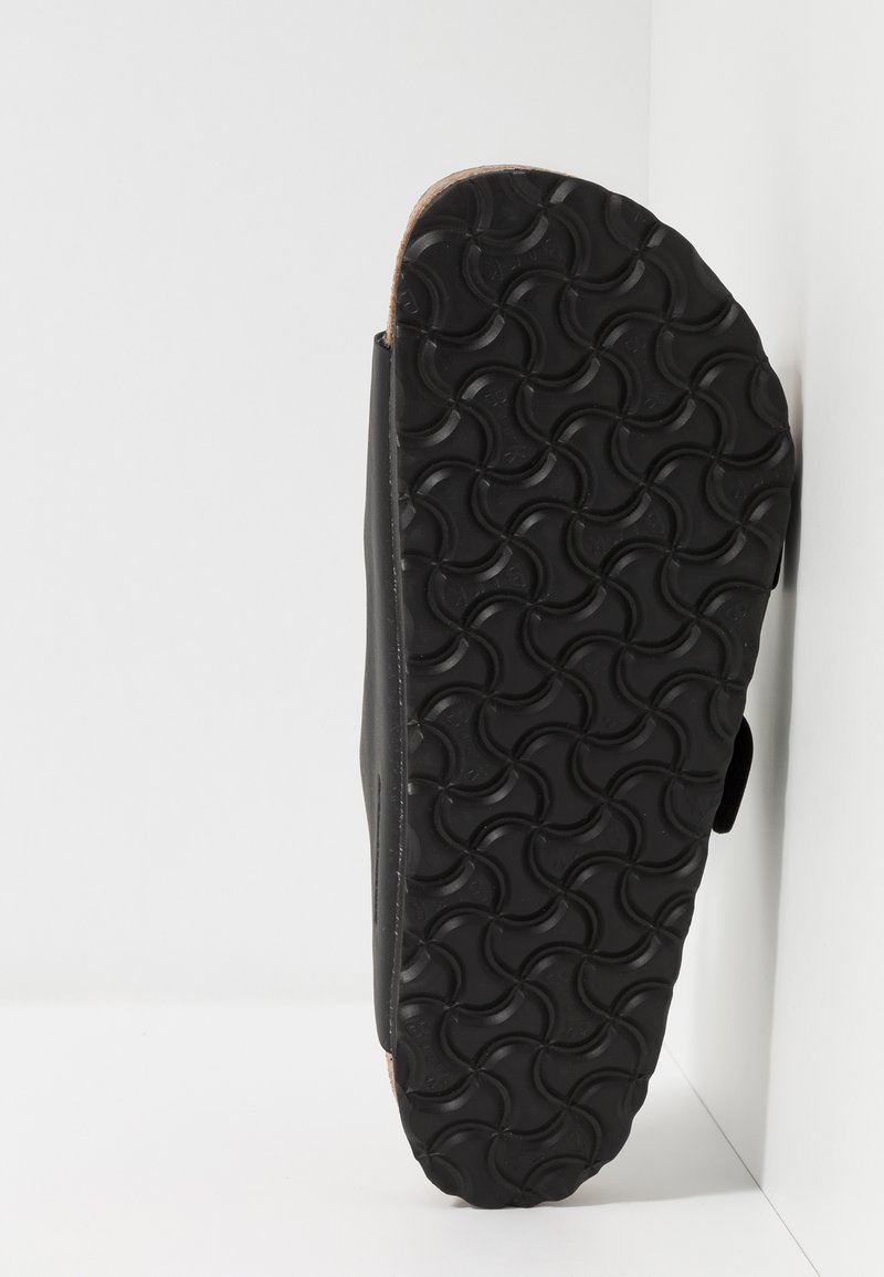 Black slide sandal featuring a textured rubber sole with circular patterns and a smooth upper. Side view shows adjustable strap for fit.