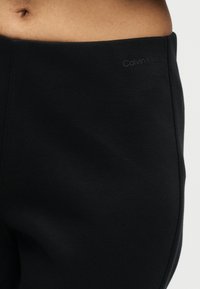 Κοντινό πλάνο ενός ατόμου που φοράει μαύρο κολάν Calvin Klein, με έμφαση στη μέση και το μερικώς εμφανιζόμενο στομάχι με ορατό αφαλό.