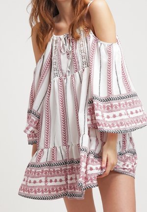 Femme portant une robe blanche à épaules dénudées avec des rayures verticales rouges et noires et des motifs floraux en bordure, tenant l'ourlet de la robe.