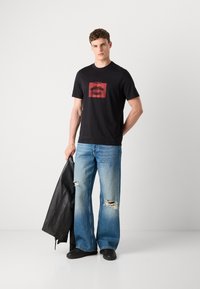 Zwarte grafische T-shirt met een design van rode lippen, gecombineerd met lichtblauwe versleten wijde jeans. Model houdt een zwarte leren jas vast.