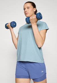 Camisa de manga corta azul claro combinada con pantalones cortos deportivos morados. Sosteniendo mancuernas azules marcadas con '3'. Tejido suave, corte holgado.