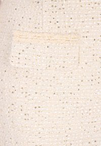 Beige rakennekankaan, jossa on pieniä kiiltäviä paljetteja ja paikallaan oleva tasku. Materiaali näyttää kudotulta ja siinä on hienostunut kiilto.