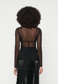 Göndör hajú nő áttetsző fekete hosszú ujjú blúzban és magas derekú fekete bőr szoknyában háttal áll egy sima háttér előtt.