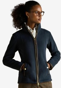 Žena s kučeravými vlasmi a okuliarmi, oblečená v námorníckej zipsovej bunde a károvaných nohaviciach, s rukami v vreckách, hľadí do strany.