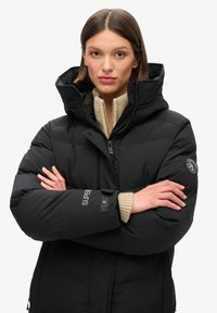 Mujer con los brazos cruzados, vistiendo una chaqueta acolchada negra con capucha y un suéter de punto beige debajo, mirando hacia adelante contra un fondo liso.