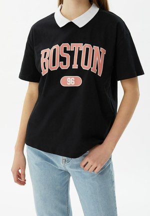 Person trägt ein schwarzes Polohemd mit kurzen Ärmeln, weißem Kragen und der Aufschrift "BOSTON 96", kombiniert mit hellblauen Jeans.