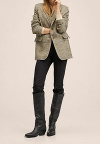 Blazer gris texturé avec boutons dorés, associé à un pantalon noir ajusté et des bottes en cuir noires montantes avec détails embossés.