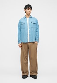 Young man wearing light blue denim shirt over white tee, loose tan pants, and black shoes, standing with hands in pockets against white background.