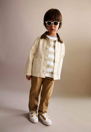 Veste beige clair avec col marron, chemise rayée blanche et beige, pantalon kaki, et baskets blanches avec des sangles jaunes. Lunettes de soleil blanches rondes.
