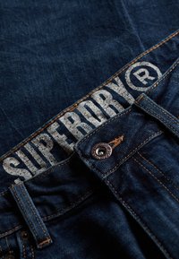 Jeans de mezclilla oscura con una cinturilla ancha que presenta el logotipo "SUPERDRY". La textura de la tela es suave con costuras contrastantes en color naranja y un botón metálico.